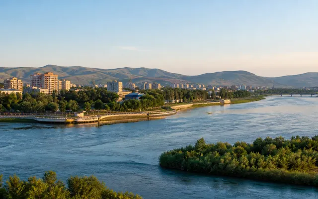 Conceptual contemporary Oskemen riverfront where broad blue water, a green embankment and low Altai foothills frame the city