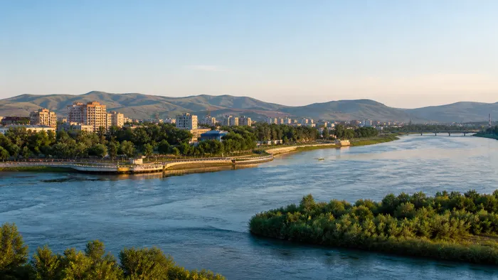 Conceptual contemporary Oskemen riverfront where broad blue water, a green embankment and low Altai foothills frame the city